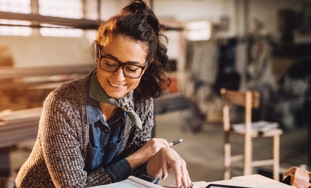 Close up view of charming smiling motivated middle aged industrial female engineer with eyeglasses working with blueprints and tablet in the workshop.